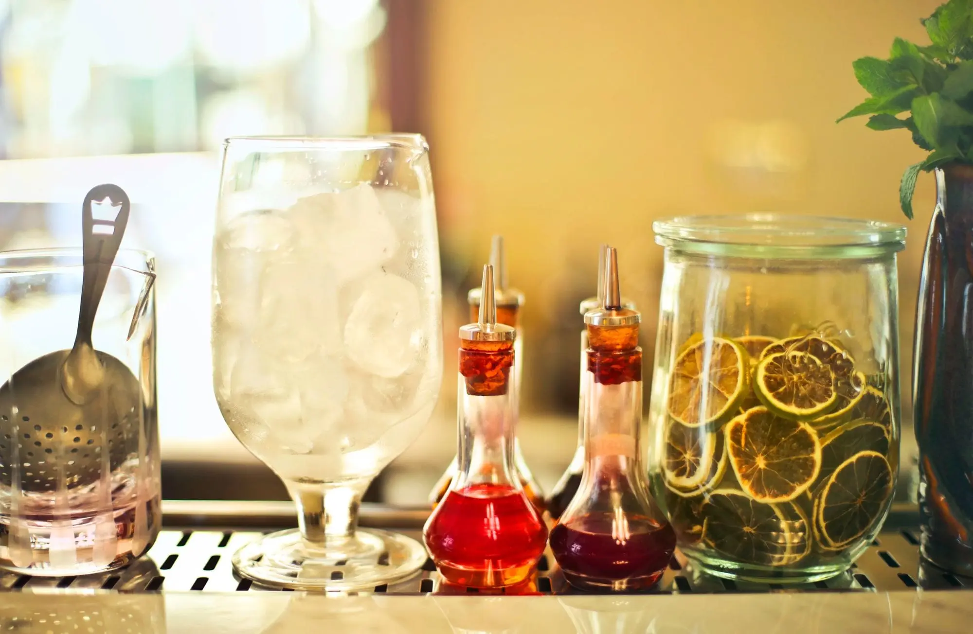 A glass full of ice on a bar counter next to bitters bottles and a jar of dried citrus