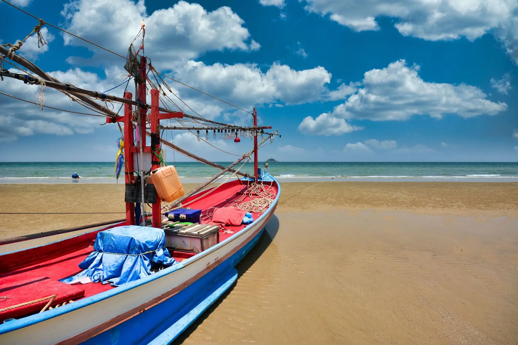A red and blue fishing boat on the sand at Hua Hin beach under a blue sky