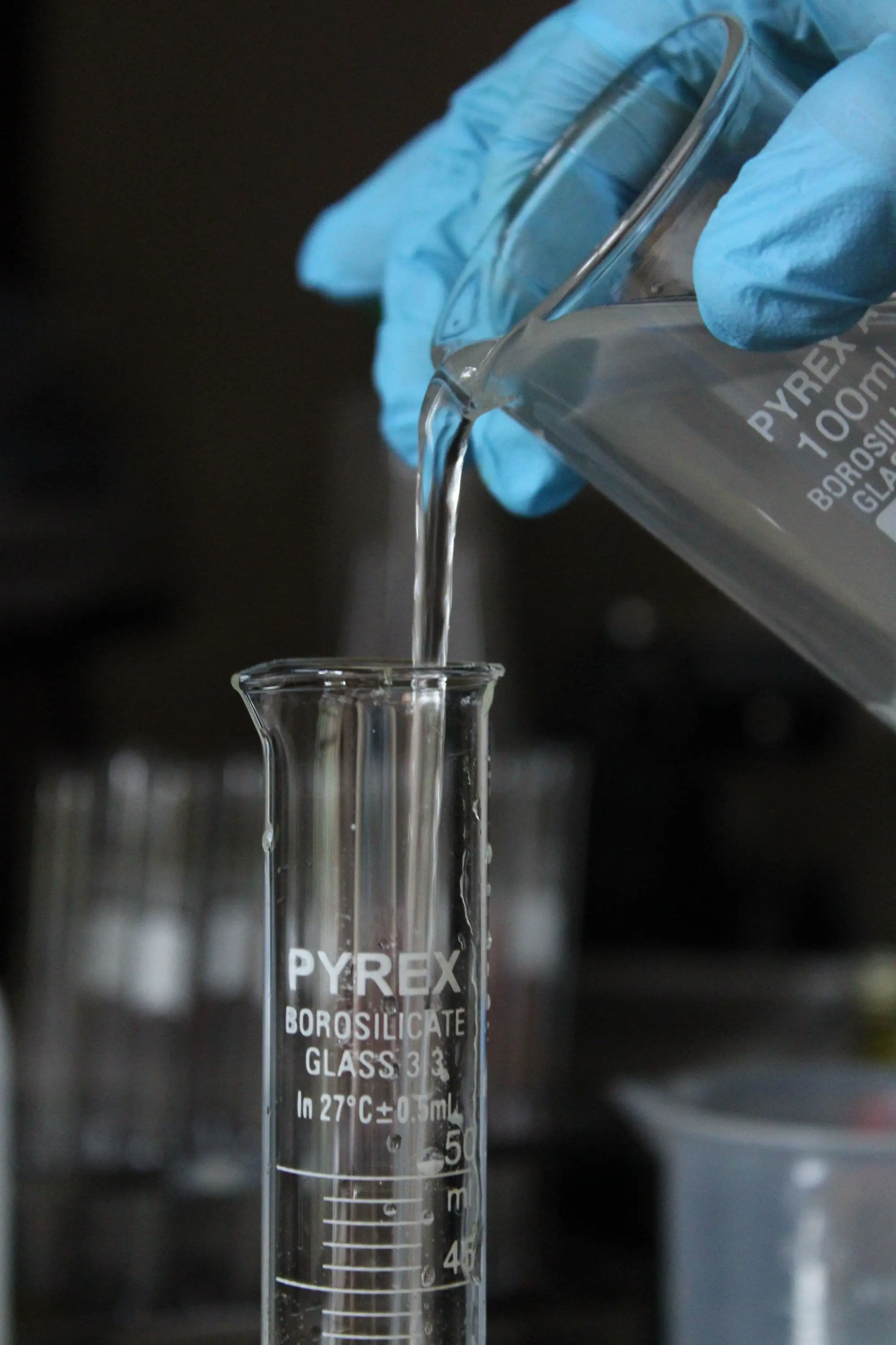Gloved hands pouring clear water from a beaker into a measuring cylinder in a laboratory